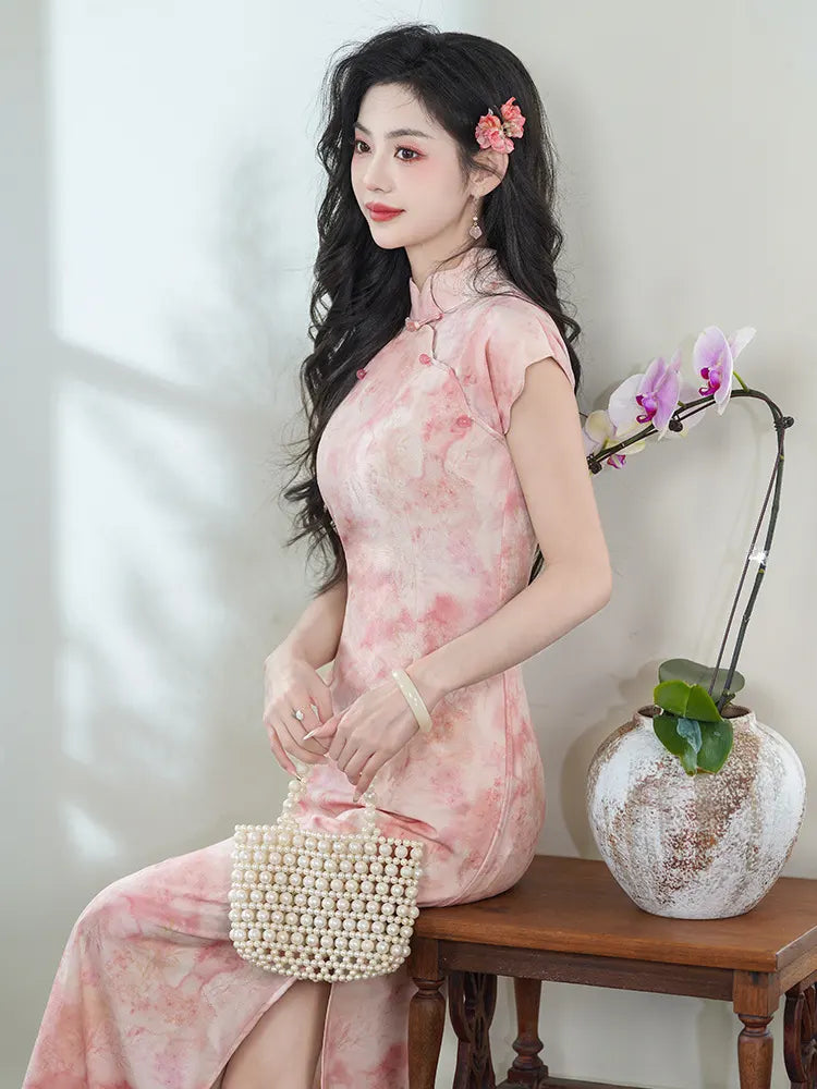 Woman in a Pink Floral Watercolor Long cheongsam  Dress sitting on a wooden bench with a vase and flowers in the background.