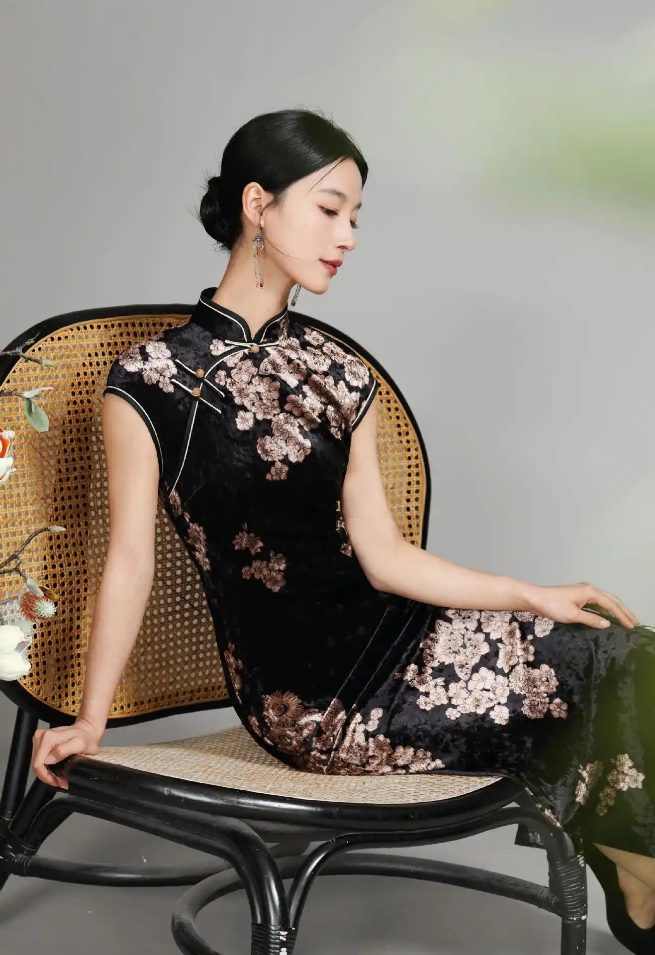 Woman in aBlack Floral Velvet Long Qipao Cheongsam Dress  sitting on a wicker chair against a gray background