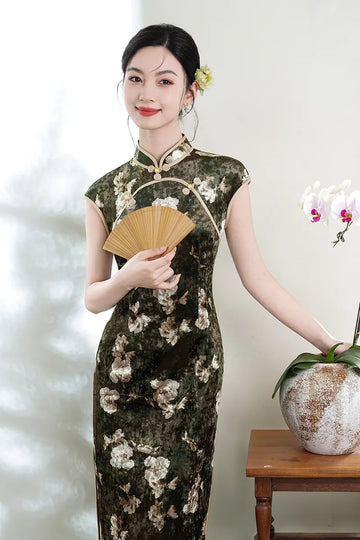 Woman in a Dark Green Foral Velvet Cap Sleeves Long Qipao Dress holding a fan, standing next to a wooden table with a vase and orchids.