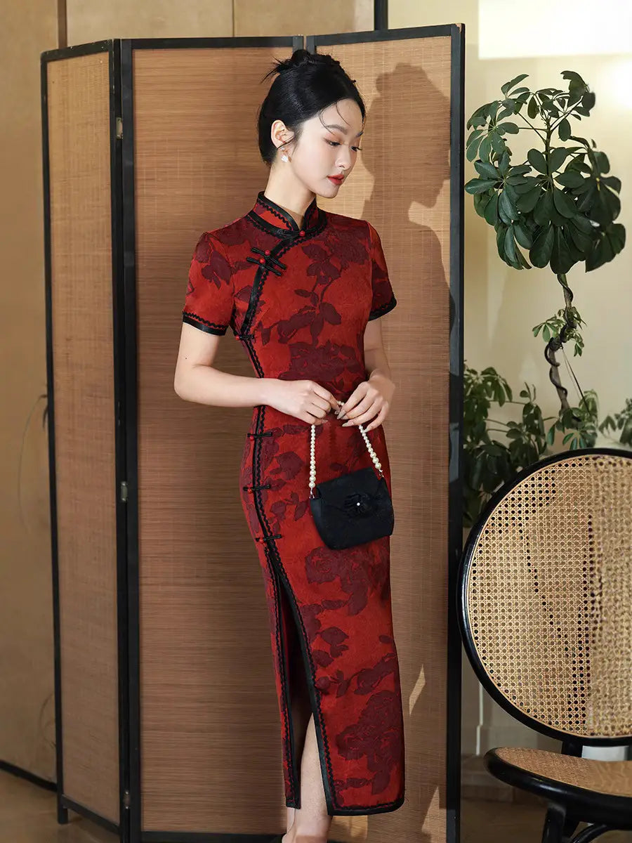 Woman in a Deep Red Floral traditional chinese outfit Qipao Cheongsam Dress with Black Trim  standing in a room with a plant and woven chair.
