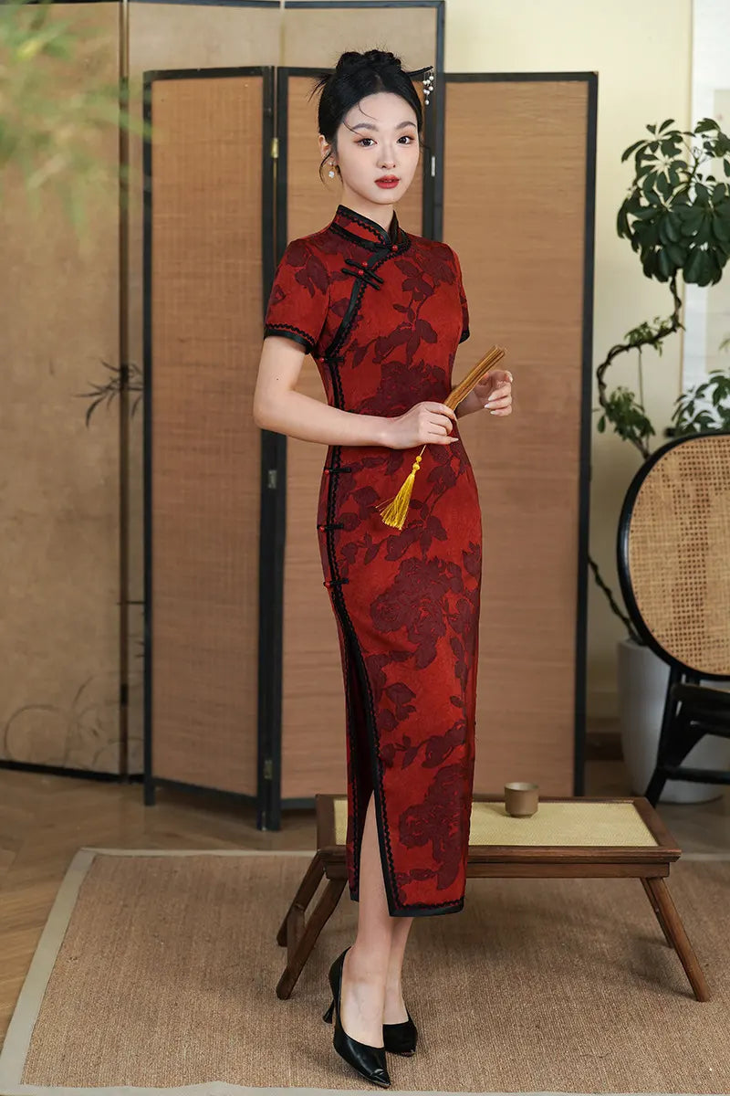 Woman in aDeep Red Floral Qipao Cheongsam Dress with Black Trim  holding hand fan in a room with wooden furniture and plants.
