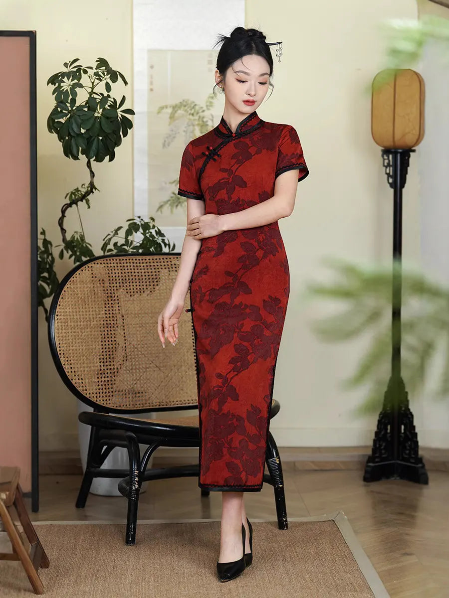 Woman in a Deep Red Floral Qipao Cheongsam Dress with Black Trim standing in a room with plants and furniture.