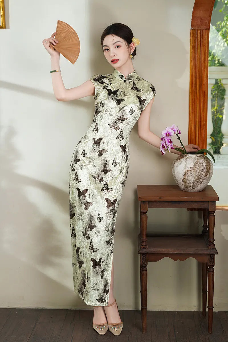 Woman in a Elegant Velvet Qipao Dress with Butterfly Print  holding a fan indoors with a wooden table and plant in the background.