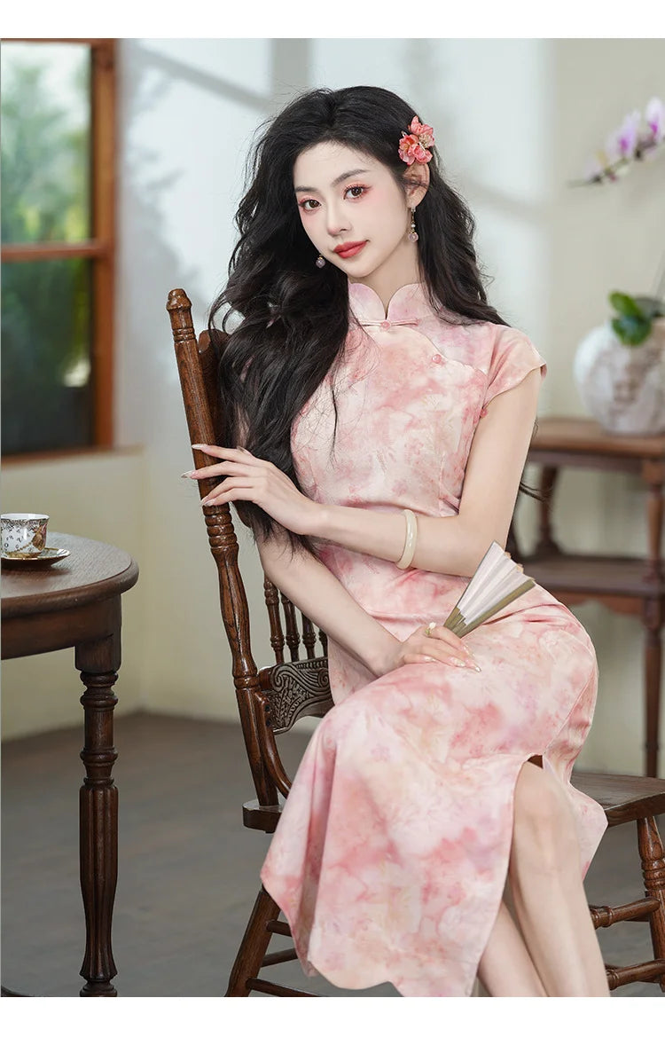 Woman in a Pink Floral Watercolor Long Qipao Dress sitting on a wooden chair indoors.