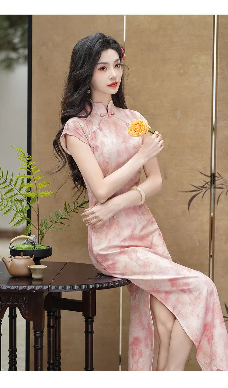 Woman in aPink Floral Watercolor Long Qipao Dress  holding a yellow flower in a room with a wooden table and plants.