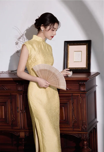 Woman in a Yellow Floral Sleeveless Midi Cheongsam Qipao Dressholding a fan in a room with wooden furniture and a framed picture.