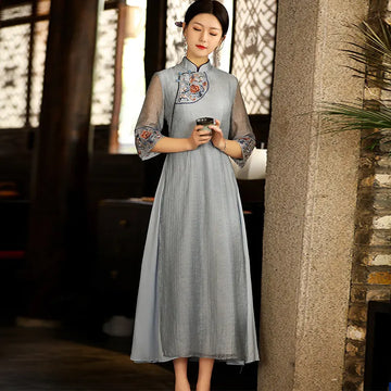 a chinese Woman in alight blue Modern A Line Loose Cheongsam Qipao Dress with Sheer Sleeve dress with floral embroidery holding a tea cup, standing against a stone wall.