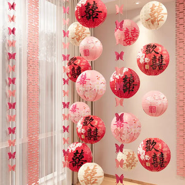Red and pink Chinese lanterns with gold double happiness characters and butterfly decorations hanging in a decorated indoor space.