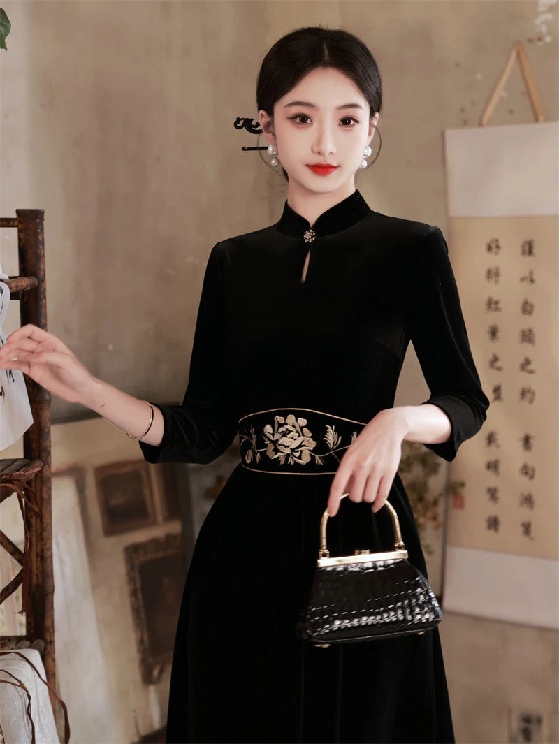 Woman in a  Black 3/4 Long Sleeves A Line Velvet Qipao Dress with   floral embroidery   with a decorative belt holding a small handbag in an indoor setting.