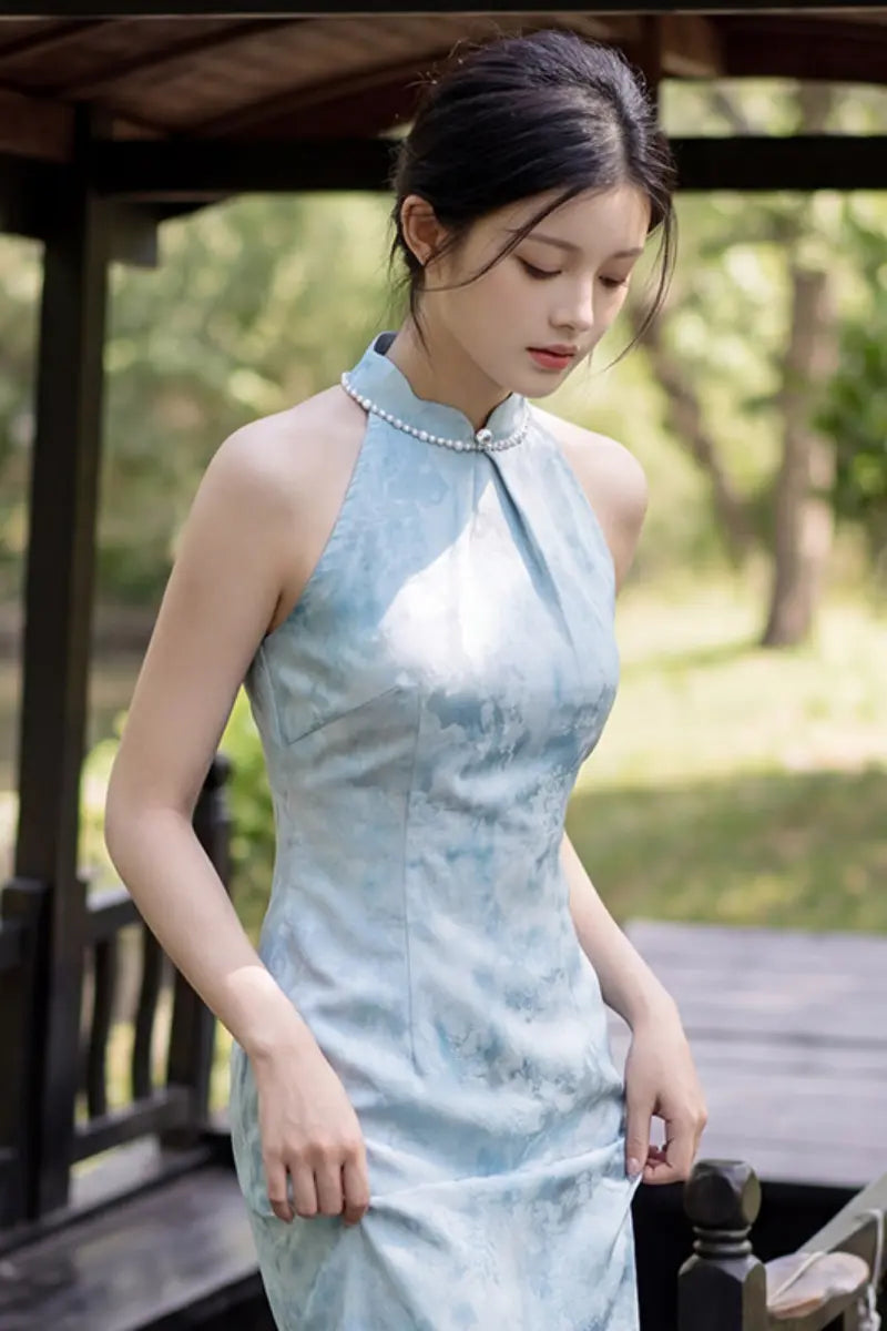 A woman wearing a sleeveless light blue and white patterned Modern Pearl-Embellished Halter-Style Qipao cheongsam dress, standing outdoors under a wooden pavilion with a blurred green garden background.