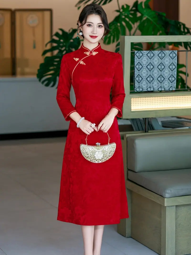 A woman stands indoors wearing a bright red long-sleeve midi cheongsam/qipao dress with gold embroidery and a small gold handbag.