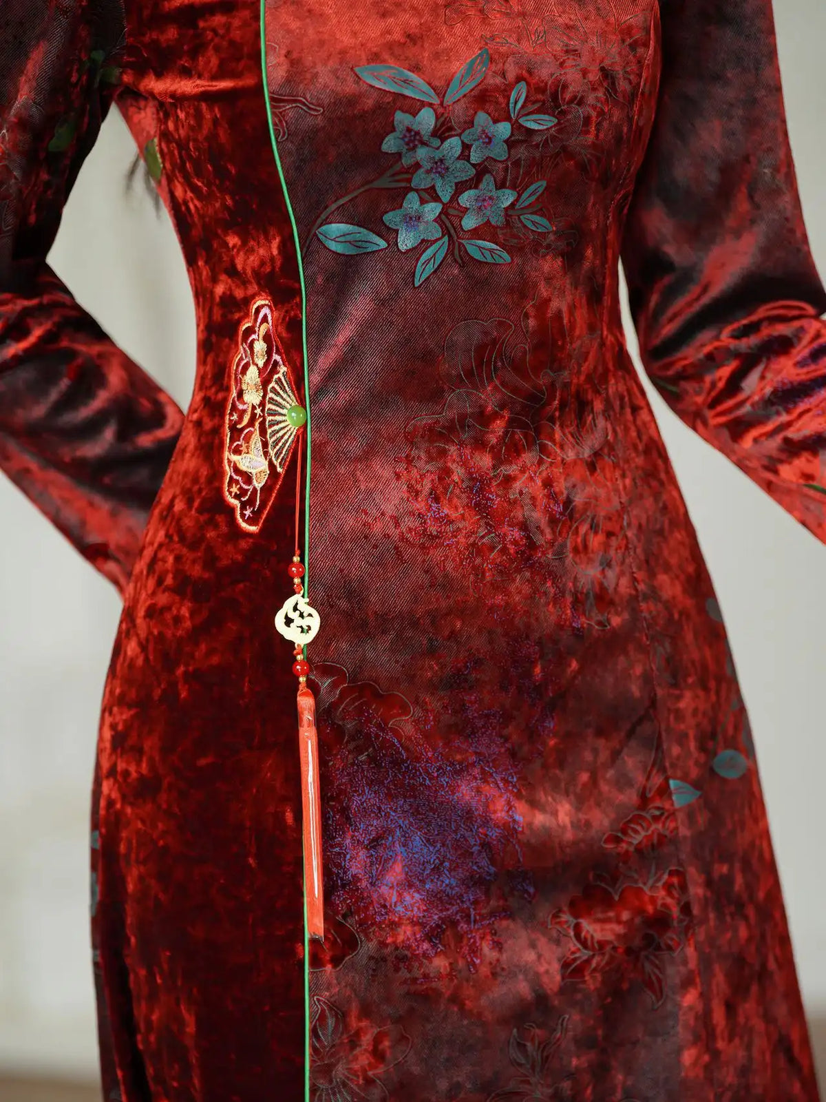 A close-up of a red velvet long-sleeve dress with blue floral embroidery on the chest, a gold embroidered motif on the hip, and a decorative tassel with a round charm hanging from a thin green string.