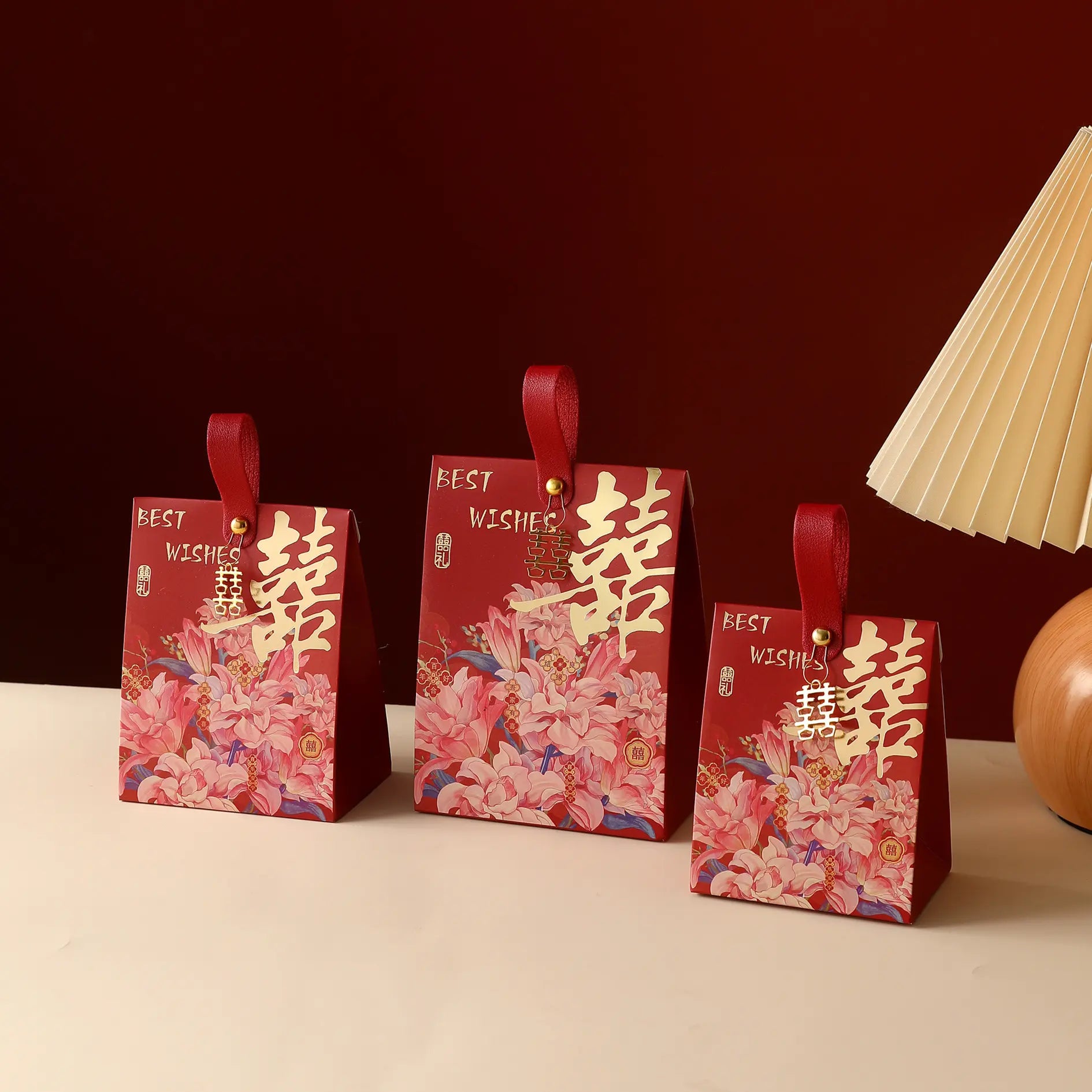 Three red Floral Double Happiness Chinese Wedding Favor Gift Boxes  with floral prints and gold Chinese characters, arranged on a light surface against a dark red background with a lamp partially visible on the right.