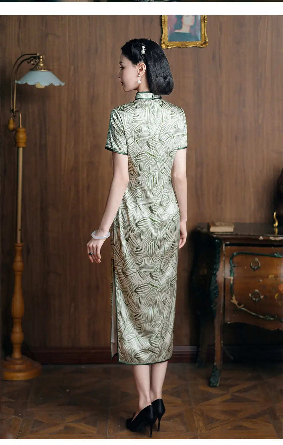 Woman in a Green Leaf Print  Chinese Silk Long Qipao Dress standing in a room with wooden walls and a lamp.