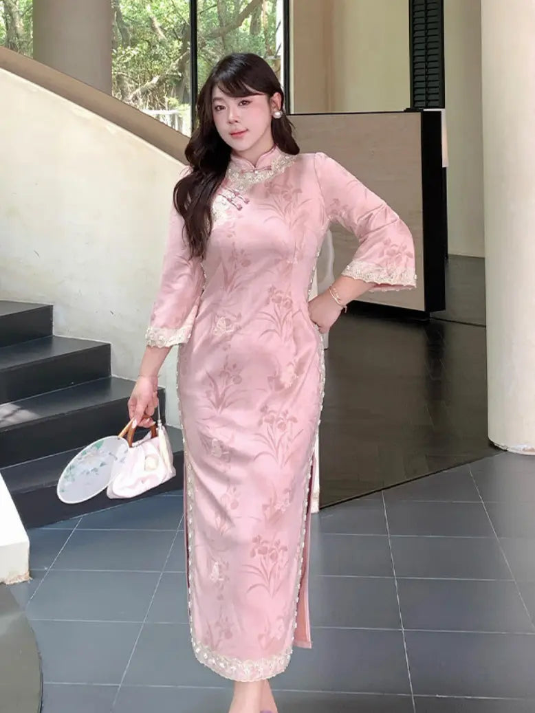 A woman wearing a light pink floral embroidered plus size cheongsam (qipao) dress with lace trim, holding a white handbag in an indoor modern setting.