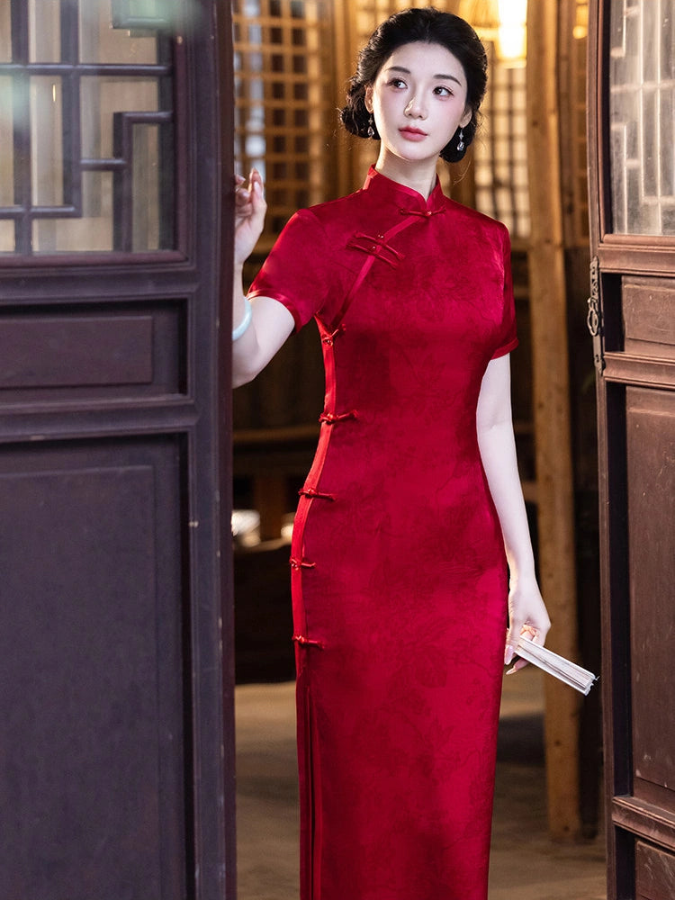 A woman wearing a vibrant red short-sleeve cheongsam (qipao) dress with traditional frog closures, standing in a doorway with wooden screens in the background.