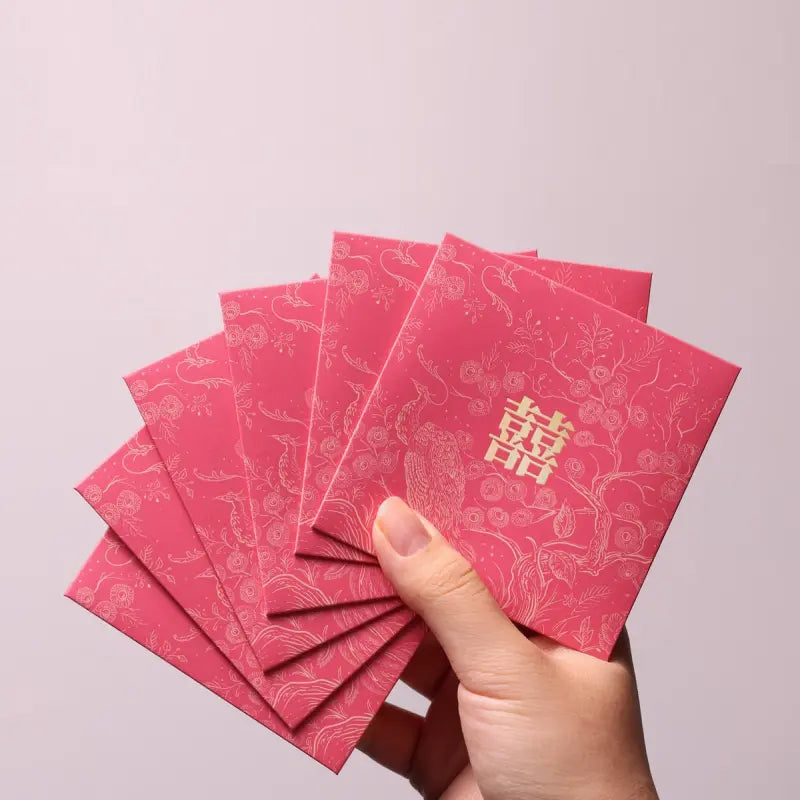 A hand holding a fan of red envelopes with gold double happiness symbols and traditional patterns, set against a plain light background.
