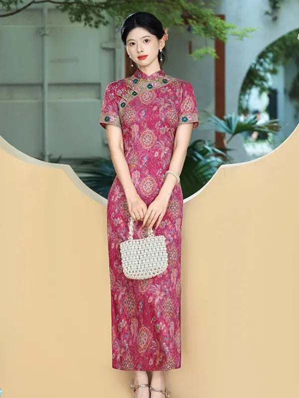 A woman stands indoors wearing a pink and red patterned cheongsam dress with short sleeves and a high collar, holding a white woven handbag. The background features a beige decorative wall and green plants.