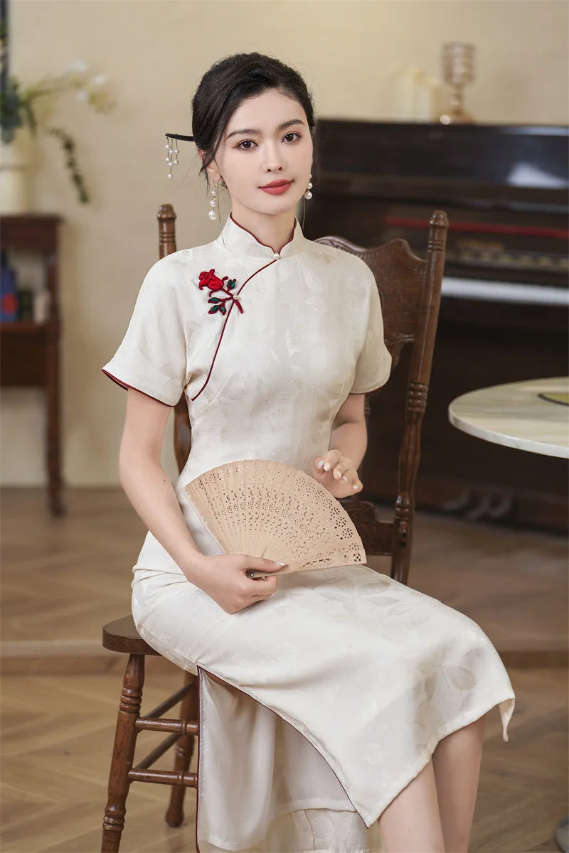 Woman in a traditional White Jacquard Qipao Cheongsam Dress With Red Trim with red floral details, holding a fan, sitting on a wooden chair.