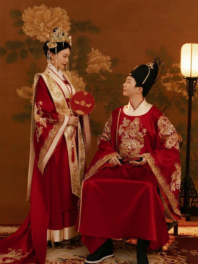 Two individuals in traditional red and gold Ming Dynasty chinese wedding hanfu attire sitting on a throne-like chair against a decorative background.