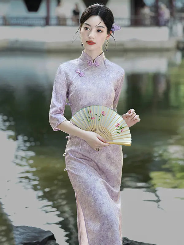 A woman wearing a light lavender floral print cheongsam (qipao) dress stands by a pond, holding a yellow folding fan with pink floral details.