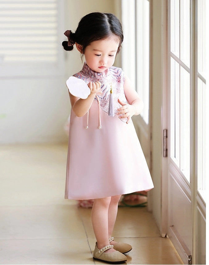 Young girl in a pink Girls' "Good Fortune" Traditional Sleeveless A-Line Jacquard Qipao dress standing in a bright room with large windows.