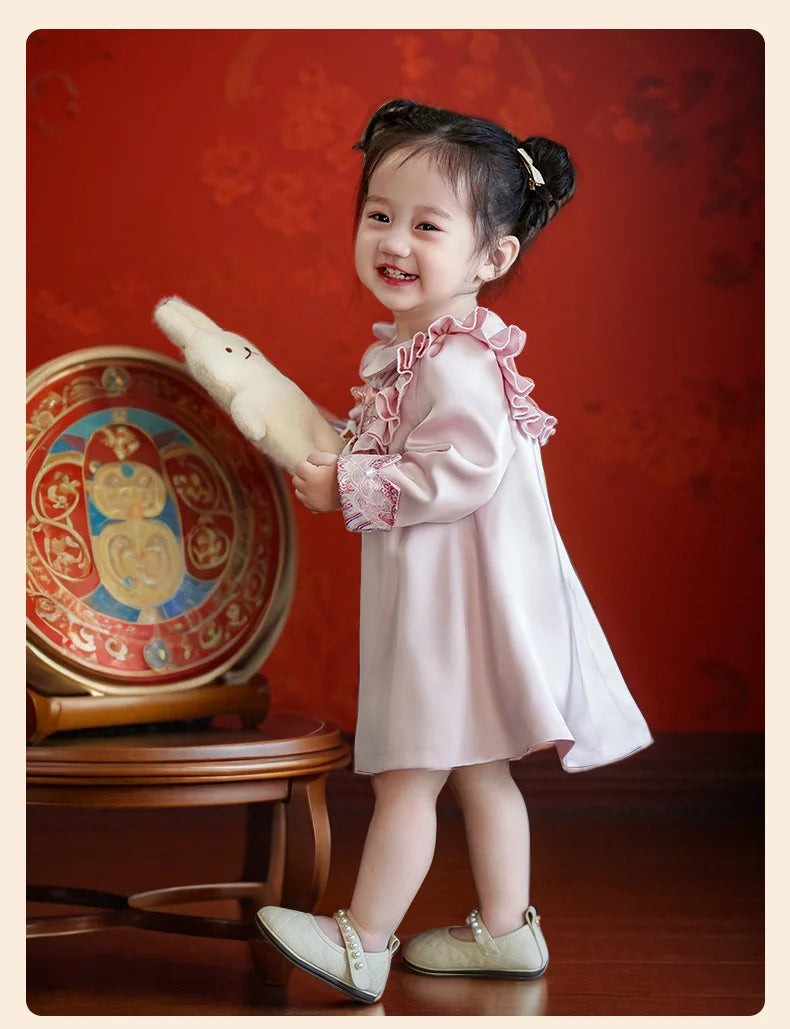 Young girl in a light pink Modern Chinese Baby Qipao Cheongsam Dress with Tassel  holding a white teddy bear against a red background