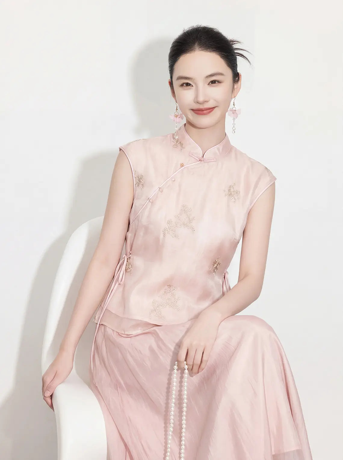Woman in a pink traditional New Chinese Style - Sleeveless Asymmetric Top & Maxi Skirt Set outfit sitting on a white chair against a white background