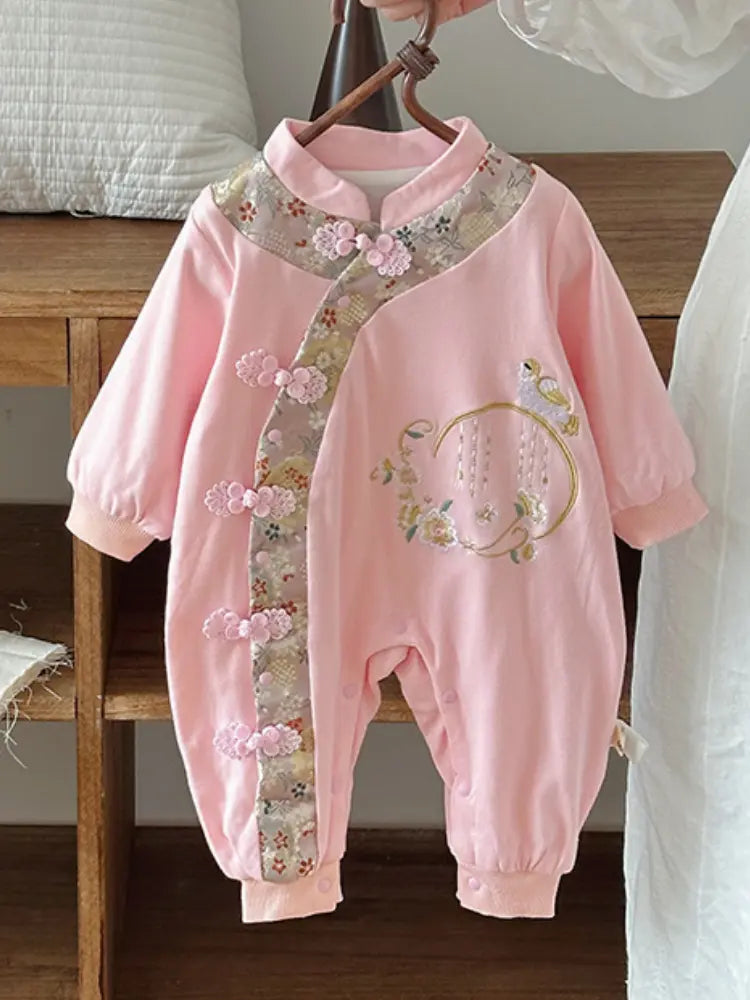 A baby girl's pink traditional-style  Chinese Embroidered Baby Girl Qipao Romper romper hanging on a wooden hanger. The romper features floral embroidery on the chest, a decorative collar, and a vertical panel of pink fabric flowers along the front closure, set against a home interior background.