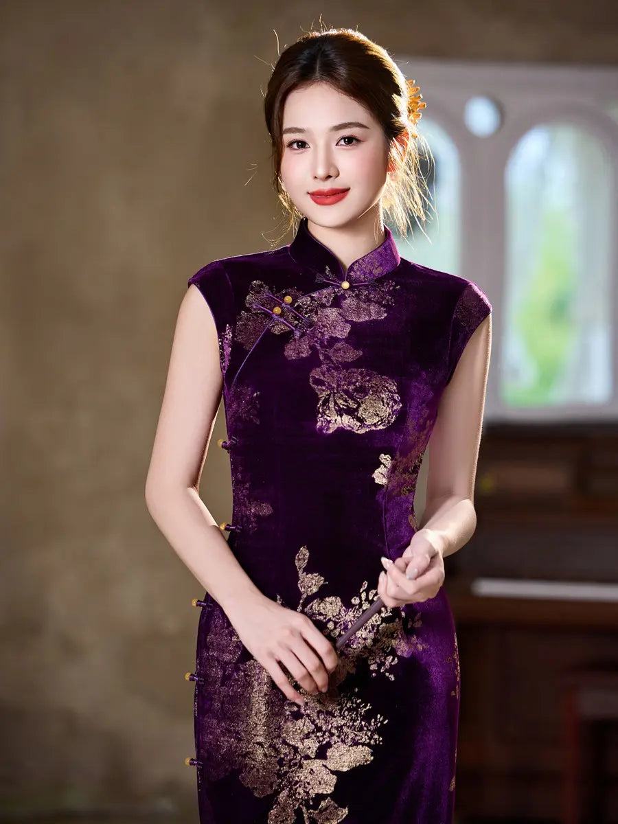 A woman wearing a sleeveless deep purple velvet cheongsam with gold floral embroidery and gold buttons, standing in a softly lit indoor setting.