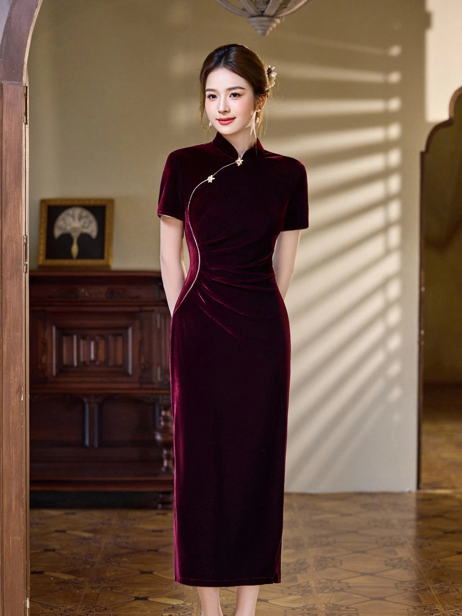 A woman wearing a dark plum red velvet cheongsam dress with a silver-toned decorative cord, standing in a warmly lit room with wooden furniture and soft window shadows.