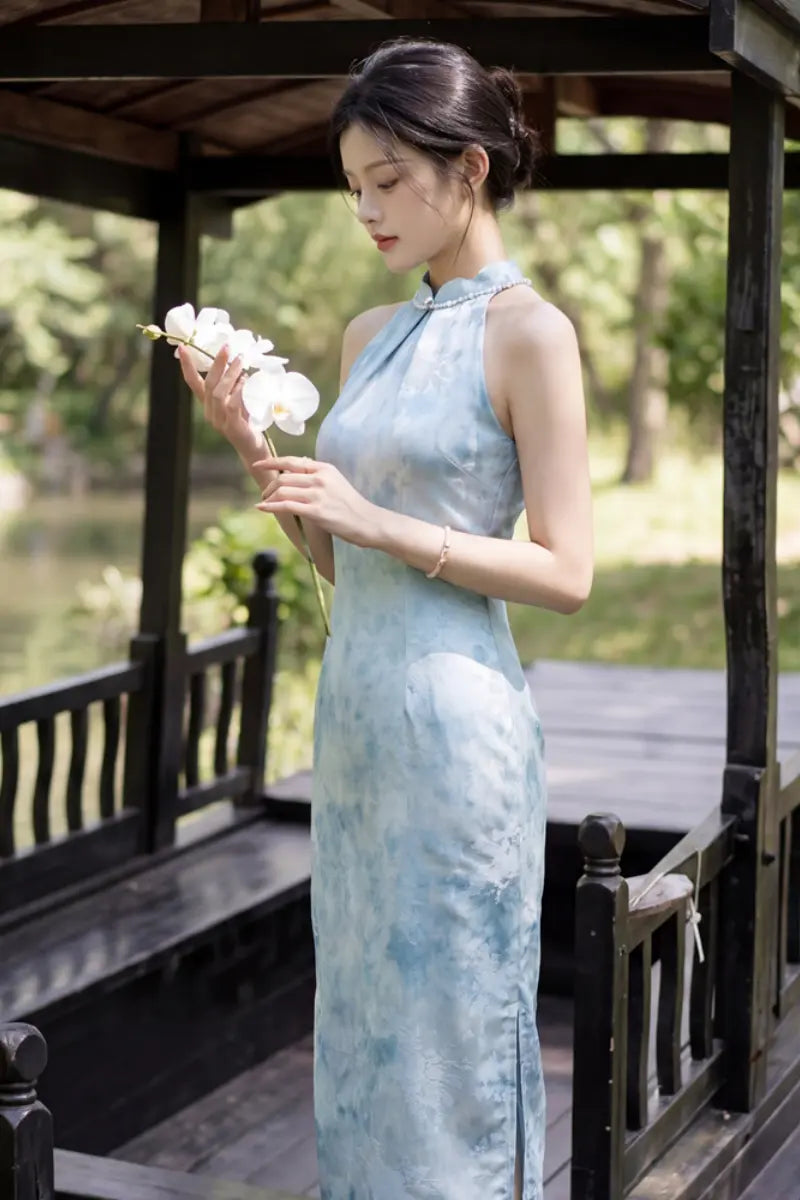 A woman wearing a light blue tie-dye Modern Pearl-Embellished Halter-Style Qipao  cheongsam dress stands in a wooden gazebo, holding a small bouquet of white flowers.