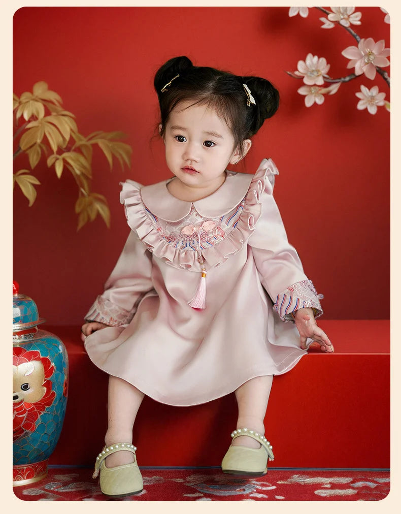 Young girl in a pink Modern Chinese Baby Qipao Cheongsam Dress with Tassel standing against a red background with floral decorations.