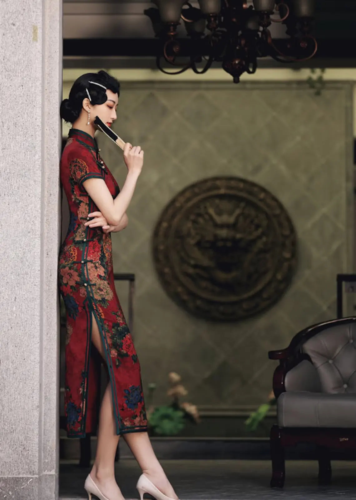 Woman in a Vintage Red Chinese Floral Qipao Cheong sam Dress standing in a room with a chandelier and decorative wall.