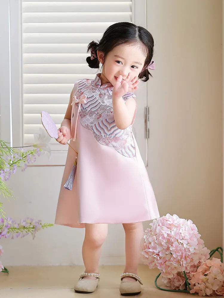 Young girl in a pink Girls' "Good Fortune" Traditional Sleeveless A-Line Jacquard Qipao dress standing indoors with flowers around her.