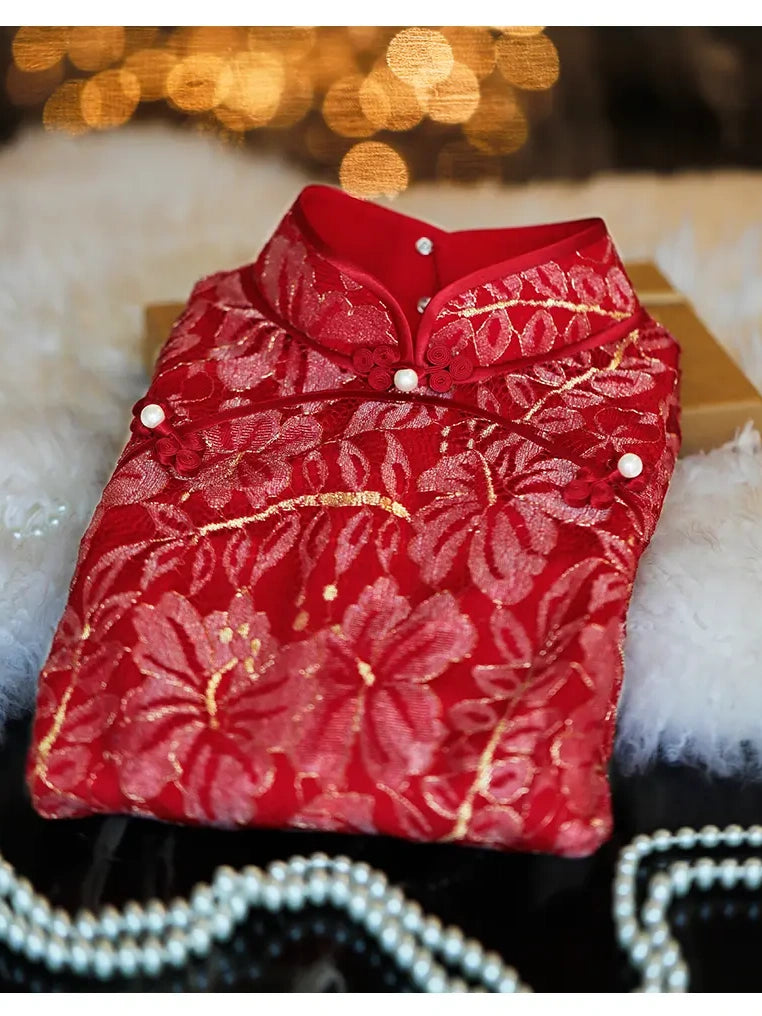 A folded red lace traditional chinese qipao dress with gold floral embroidery and pearl-like buttons, displayed on a soft white faux-fur surface with warm blurred lights in the background.
