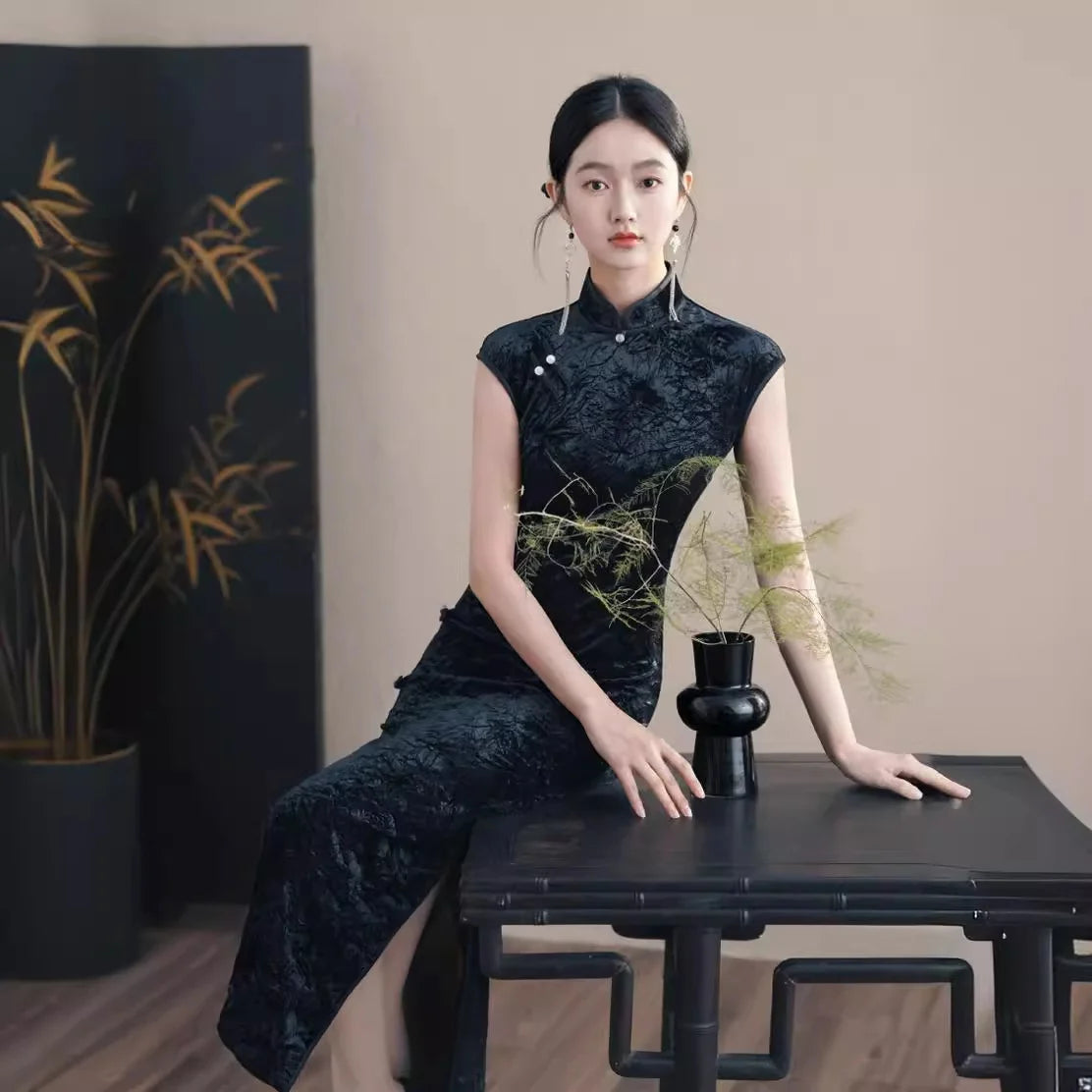 Woman in a Black Floral Velvet Qipao Cheongsam  traditional outfit sitting at a table with a vase and plant in a minimalistic setting.