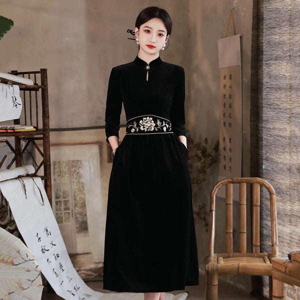 Woman wearing a Black 3/4 Long Sleeves A Line Velvet Qipao Dress with   floral embroidery in a room with a chair and shelves.