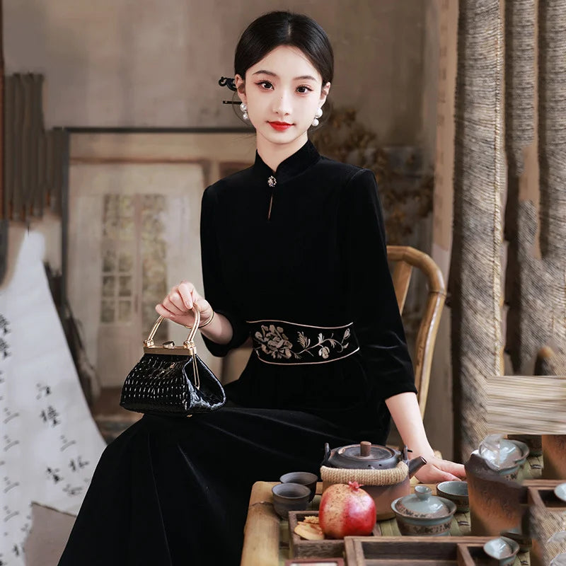 Woman in a traditional  Black 3/4 Long Sleeves A Line Velvet Qipao Dress with   floral embroidery  outfit holding a small black handbag, sitting at a tea table.