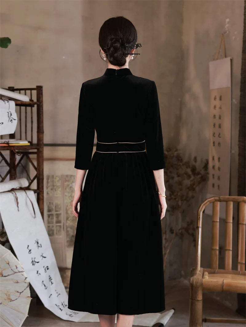 Woman wearing a  Black 3/4 Long Sleeves A Line Velvet Qipao Dress with   floral embroidery  standing in a room with a textured wall and wooden chair.