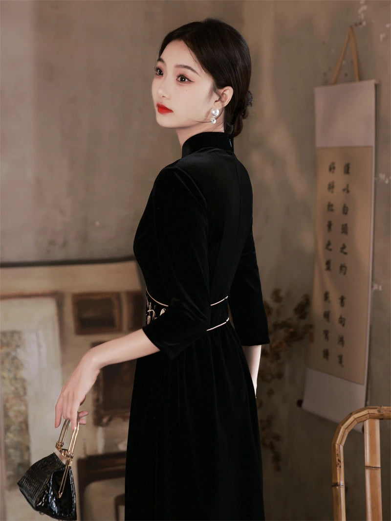 Woman in a  Black 3/4 Long Sleeves A Line Velvet Qipao Dress with   floral embroidery   holding a handbag in an indoor setting with a textured wall and framed picture.
