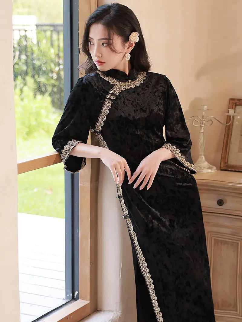 A woman wearing a black velvet cheongsam-style dress with gold lace trim, standing by a window in a warm indoor setting.