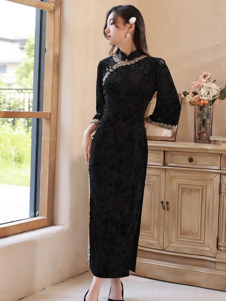 A woman wearing a black long-sleeved lace cheongsam dress with gold embroidery on the collar and cuffs, standing in a room with a wooden cabinet and window.
