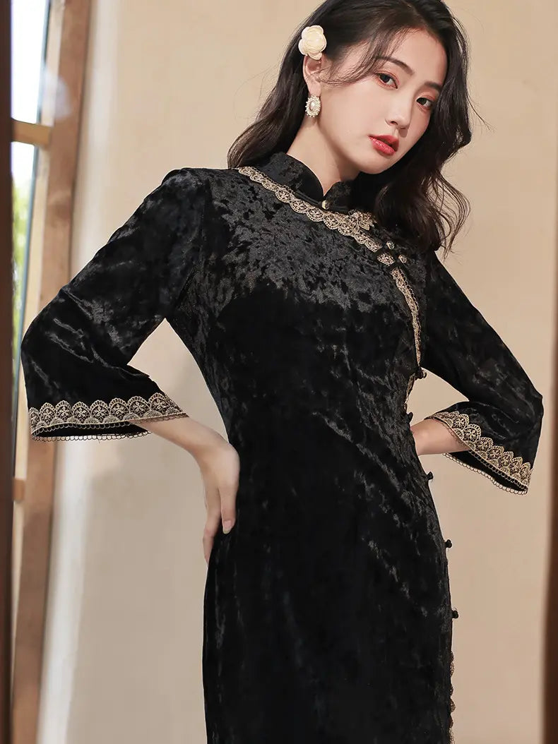 A woman wearing a black velvet cheongsam-style dress with gold lace trim and patterned cuffs, posed indoors against a soft beige background.