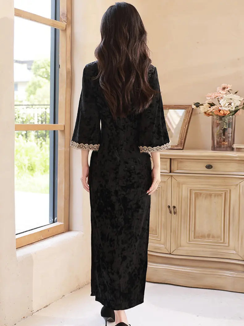 A woman viewed from behind wearing a long black textured dress with wide three-quarter sleeves and lace trim at the cuffs, standing in a bright room with a window and wooden cabinet.