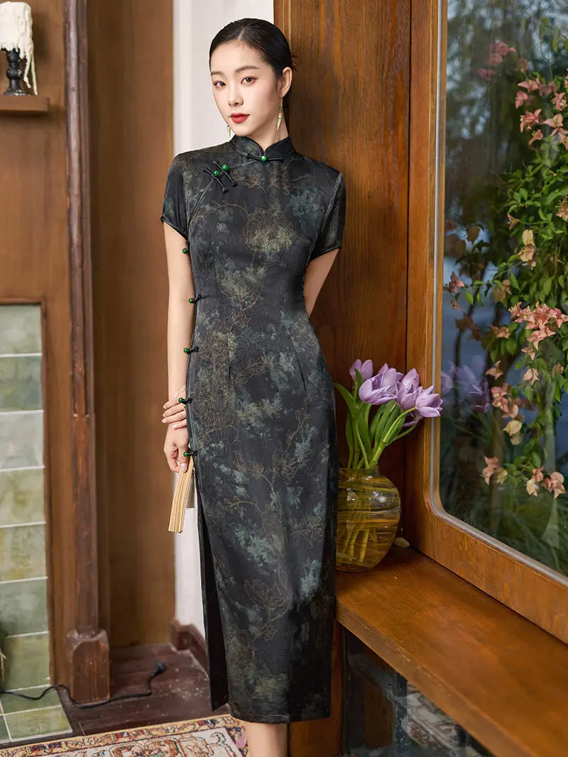 Woman in a Black & Deep Emeral Jacquard Satin Long Qipao cheogsam With Green Buttons  standing indoors next to a window with flowers.