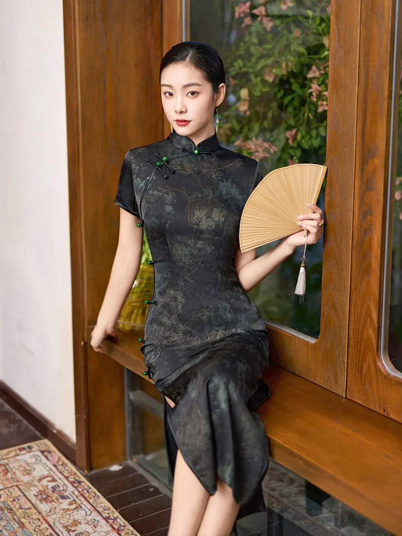 Woman in a traditional chinese Black & Deep Emeral Jacquard Satin Long Qipao With Green Buttons dress holding a fan indoors.