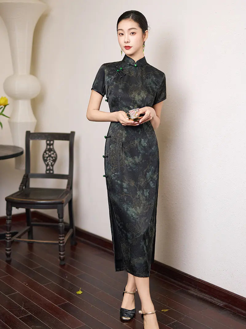 Woman wearing a Black & Deep Emeral Jacquard Satin Long Qipao With Green Buttons  standing in a room with a chair and table.
