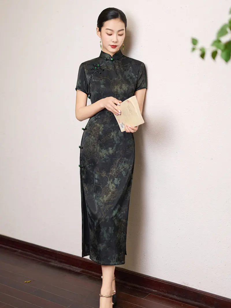Woman wearing a Black & Deep Emeral Jacquard Satin Long Qipao With Green Buttons holding a clutch against a white wall.