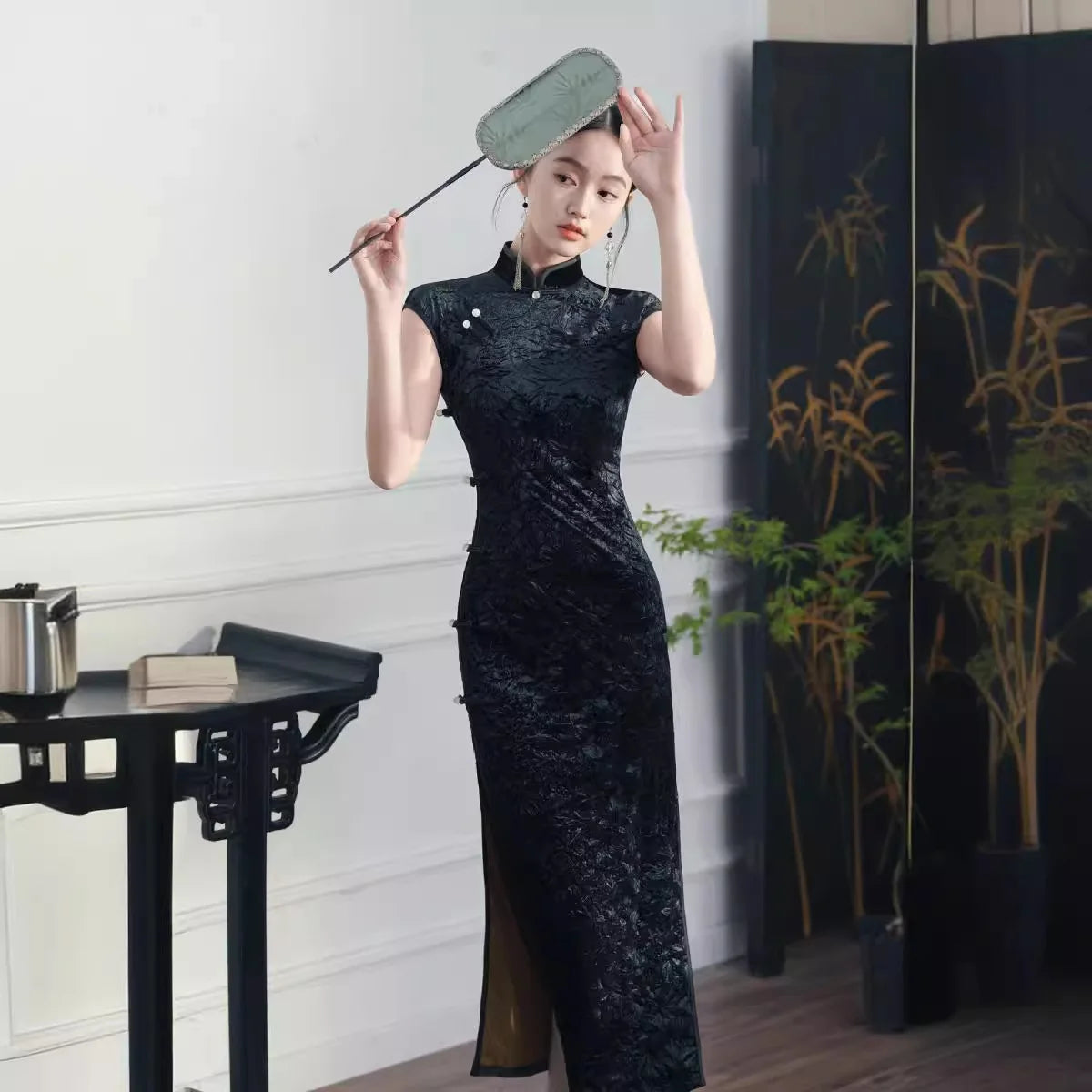 Woman in a  traditional Black Floral Velvet Qipao Cheongsam Dress  holding a fan in a room with a black screen and plant.